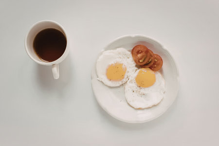 A plate with two fried eggs and sliced tomatoes next to a cup of coffee on a white table.