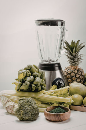 A blender surrounded by various fruits and vegetables, including broccoli, celery, pineapple, and...