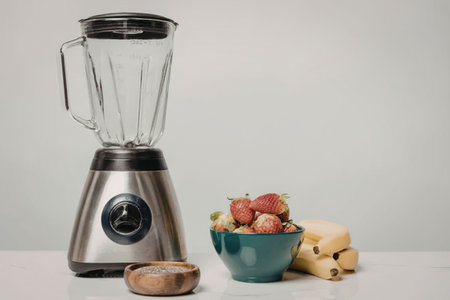 A blender placed next to a bowl of strawberries, a few bananas, and a wooden lid on a white surface.