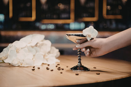 A hand holding a cocktail glass on a wooden table with coffee beans scattered around.
