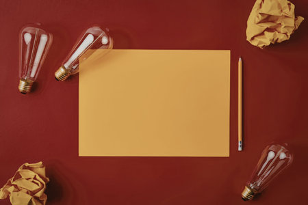 A vibrant workspace setup featuring a blank orange paper, surrounded by light bulbs and crumpled...