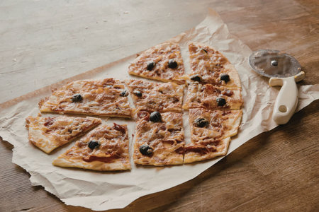 A mouth-watering homemade pizza cut into slices on a wooden table with a pizza cutter.