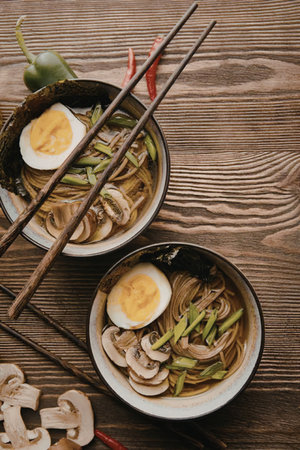 Two bowls of noodles with boiled eggs and mushrooms on a wooden table.