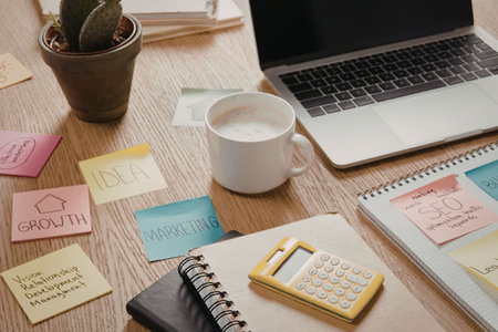 A wooden desk with a laptop, coffee cup, plant, calculator, notebook, and colorful sticky notes.