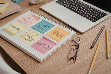 A wooden desk with a laptop, spiral-bound notebook with sticky notes, glasses, and pencils.