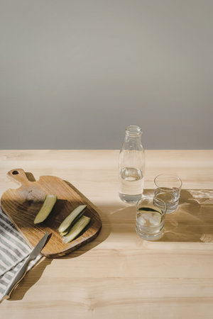 A wooden cutting board with sliced cucumbers, a knife, and a glass bottle with two glasses of water...