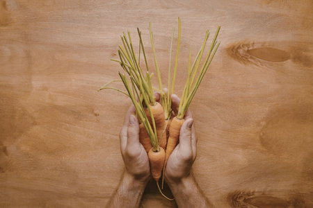 A pair of hands holding a bunch of fresh carrots with green tops on a wooden surface