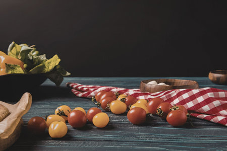 A vibrant display of cherry tomatoes on a rustic wooden table with a red and white checkered cloth, accompanied by a bowl of greens and a wooden spoon.の写真素材