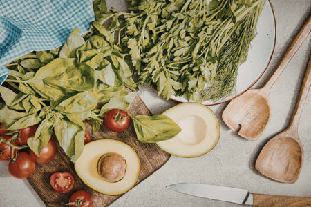 A kitchen counter with fresh vegetables and utensils, including cherry tomatoes, basil, avocado, and...
