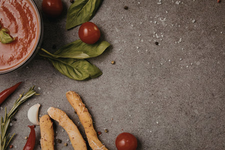 A bowl of fresh tomato sauce with breadsticks, tomatoes, and herbs on a gray surface