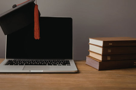 A graduation cap sits on top of a laptop next to a stack of books on a wooden desk