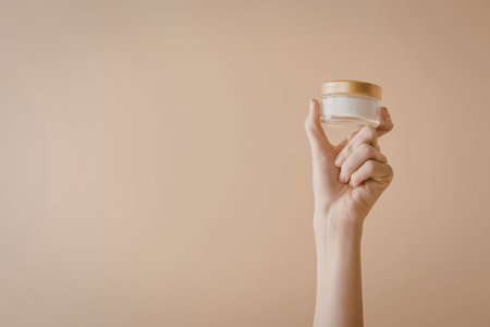 A hand holding a small jar of cream against a beige background