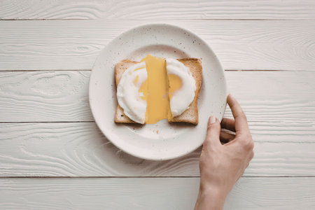 A hand holding a white plate with toasted bread topped with a poached egg on a white wooden table.