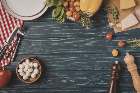 A wooden table with various fresh ingredients and kitchen utensils, including tomatoes, cheese,...