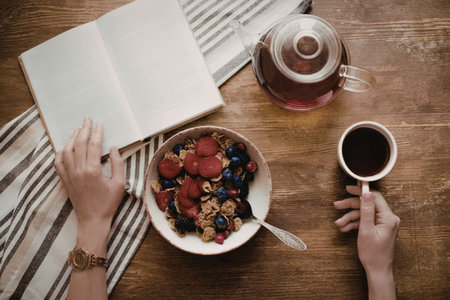 An overhead shot of a person's hands holding a cup of tea and a fork, with a bowl of cereal and an...