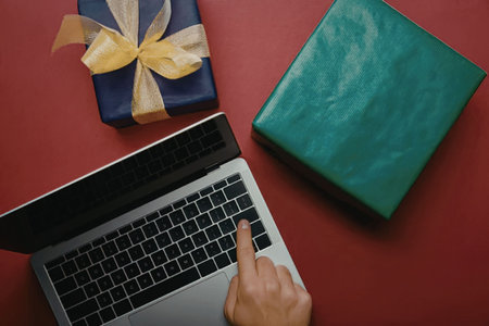 A laptop with a hand on the keyboard next to two gift boxes on a red surface