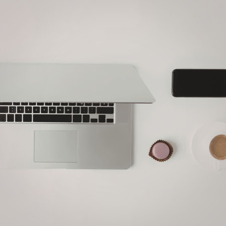 A modern workspace setup featuring a laptop, smartphone, cup of coffee, and a macaron on a clean...