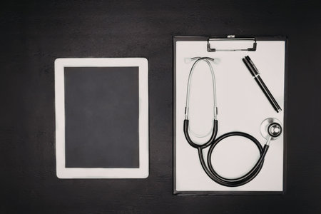 A stethoscope, pen on a clipboard and a tablet on a black background
