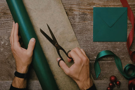 A person's hands are shown cutting green wrapping paper and preparing a gift with ribbon and red berries on a wooden surface.の写真素材