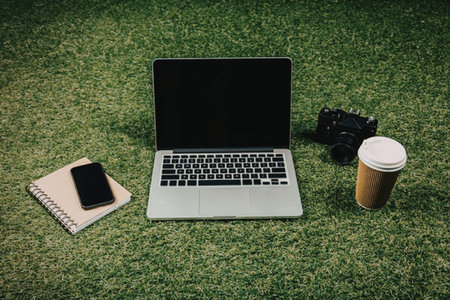 A laptop, notebook, camera and coffee cup on green grass