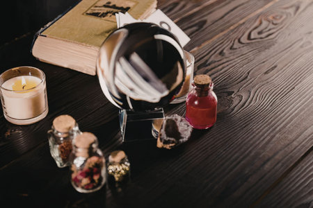 A dark wooden table holds various items including jars with potions and herbs, a lit candle, and an...