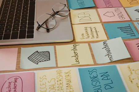 A laptop and glasses sit on a desk covered with colorful sticky notes containing business-related...