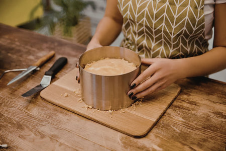 A woman in a green and white patterned top is preparing a cake on a wooden cutting board with a...