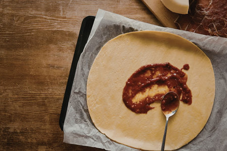 A pizza dough being prepared with tomato sauce on a wooden table.