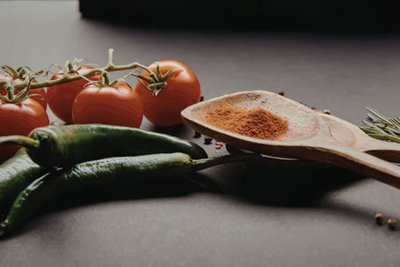 A wooden spoon with red spice, green peppers, and tomatoes on a dark surface