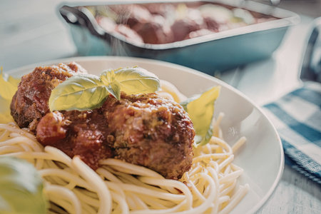 Spaghetti with meatballs and tomato sauce is served on a white plate garnished with fresh basil leaves on a blue and white checkered tablecloth.の写真素材