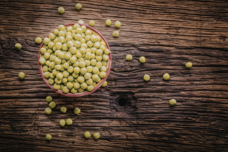 Fresh green peas fill a small red bowl on a rustic wooden table with scattered peas surrounding it,...