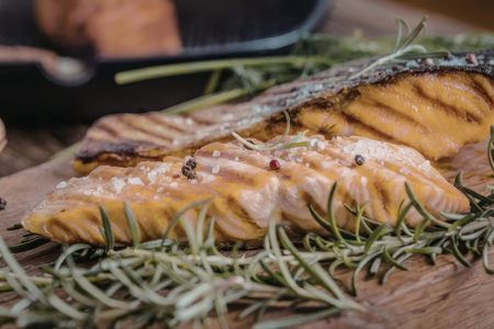 Grilled salmon fillets seasoned with rosemary and salt are placed on a wooden cutting board surrounded by fresh herbs.の写真素材