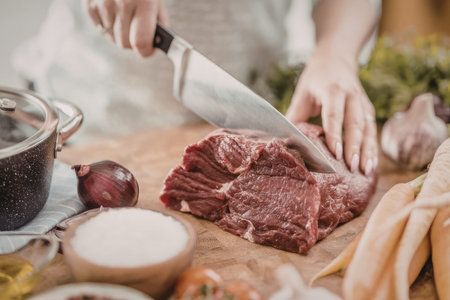 Person wearing white shirt skillfully chops raw meat on a wooden cutting board surrounded by various...