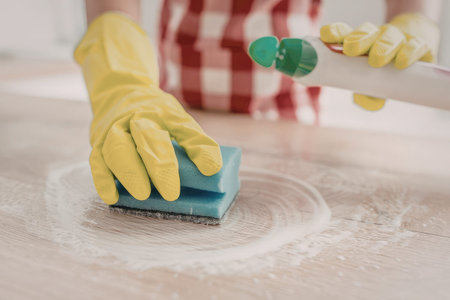 Person wearing yellow rubber gloves wipes a wooden surface with a blue sponge and white cleaning solution to maintain cleanliness and hygiene.の写真素材