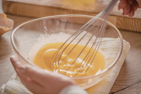 Person holding a clear glass bowl containing a yellow egg mixture being whisked with a metal whisk...