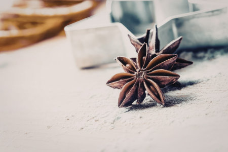 Star anise lies on a light textured surface with blurred baking utensils and ingredients in the...