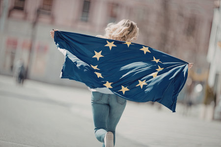 Young woman with blonde hair runs down a city street with european union flag waving in the wind.