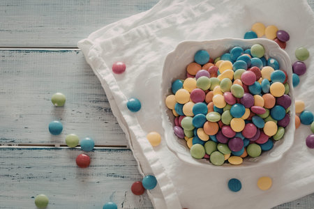 Colorful candy coated chocolate candies spill out of a white bowl onto a white cloth on a distressed...