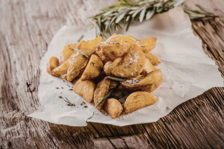 Crispy fried potato wedges sprinkled with coarse salt and rosemary on a rustic wooden surface creating a delicious and savory snack experience.の写真素材