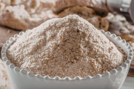 Bread crumbs fill a small white bowl with freshly baked bread visible in the blurred background,...