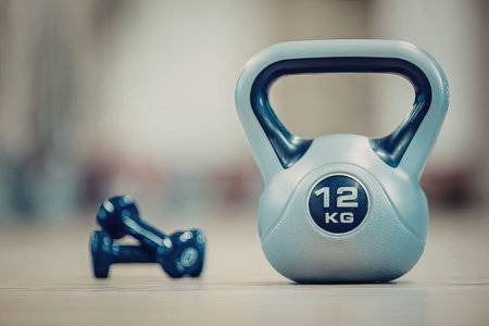Kettlebell weighing 12 kg sits on a wooden floor alongside two small blue dumbbells in a blurred gym...