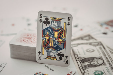 King of clubs playing card stands upright amidst scattered cards and dollar bills on a white...