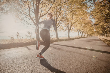 Young woman runs on a scenic path surrounded by trees with autumn leaves, enjoying a sunny day and a...