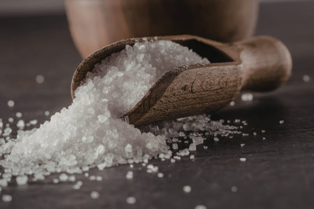 Coarse sea salt spills from a wooden scoop onto a dark surface, showcasing its texture and crystals...