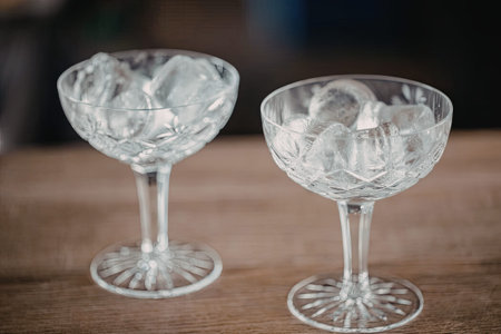 Crystal clear elegant cocktail glasses filled with ice sit on a wooden table ready to be filled with...