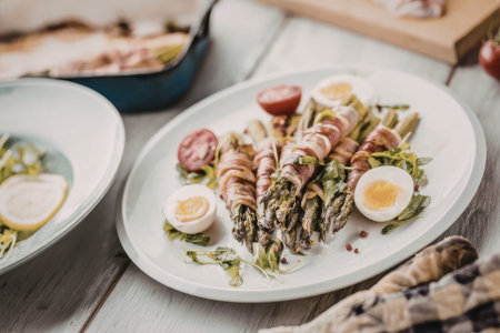 Asparagus wrapped in bacon served with boiled egg halves and fresh herbs on a white oval plate symbolizing a healthy gourmet meal.の写真素材