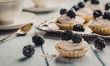 Sweet pastry tartlets filled with cream and topped with blackberries on a rustic white wooden table with a delicate dessert setting.の写真素材