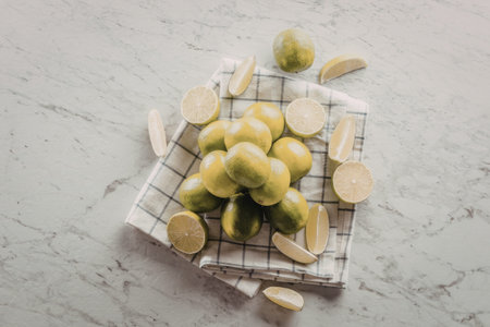 Fresh limes and lime slices arranged on a checkered cloth placed on a marble countertop, showcasing...