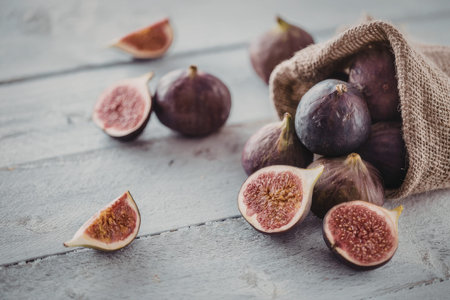 Fresh ripe purple figs spill out of burlap sack on rustic wooden table with some cut open displaying...