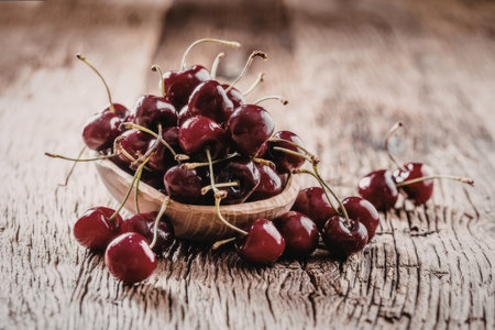 Fresh ripe cherries overflow from a small wooden bowl, scattered on a rustic wooden surface,...